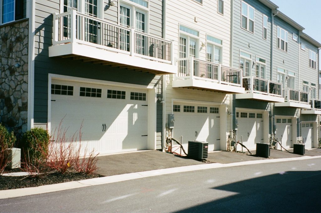 A row of modern townhomes with beige and gray exteriors. Each unit has a white garage door, balconies with railings above the garages, and air conditioning units beside the driveways. The street in front is clean and vacant, with minimal landscaping.