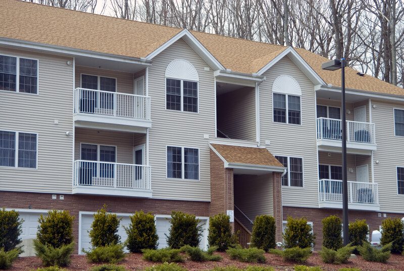 A two-story apartment building with beige siding and a brown roof features balconies with white railings. Bushes and small trees are planted in front of the building, and a streetlight is visible on the right side. The design allows for easy apartment cleaning, ensuring every unit remains pristine.