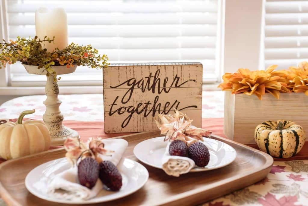 A cozy dining table setup for Thanksgiving featuring a "gather together" sign, decorative pumpkins, and dried corn on white plates. The table is adorned with a candle centerpiece and orange flowers, creating a warm, autumnal ambiance to help you prepare your home for the holiday season.