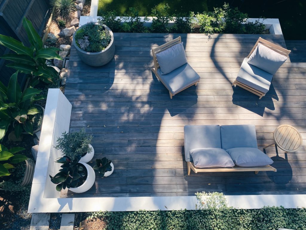 Aerial view of a clean deck featuring a wooden patio with cushioned chairs and a loveseat. The patio, perfect for the spring season, is adorned with potted plants and bathed in a mix of sunlight and shadow. Surrounding the patio is greenery, adding a natural ambiance to the cozy outdoor space.