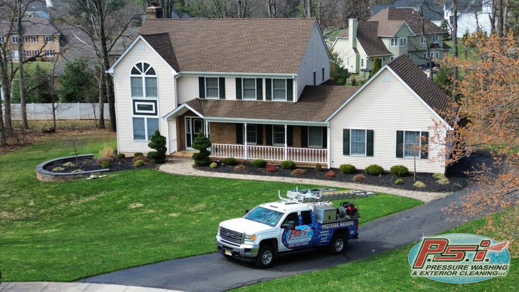 Two-story beige house with brown roof and a wraparound porch; a pressure-washing truck is parked on the street in front.