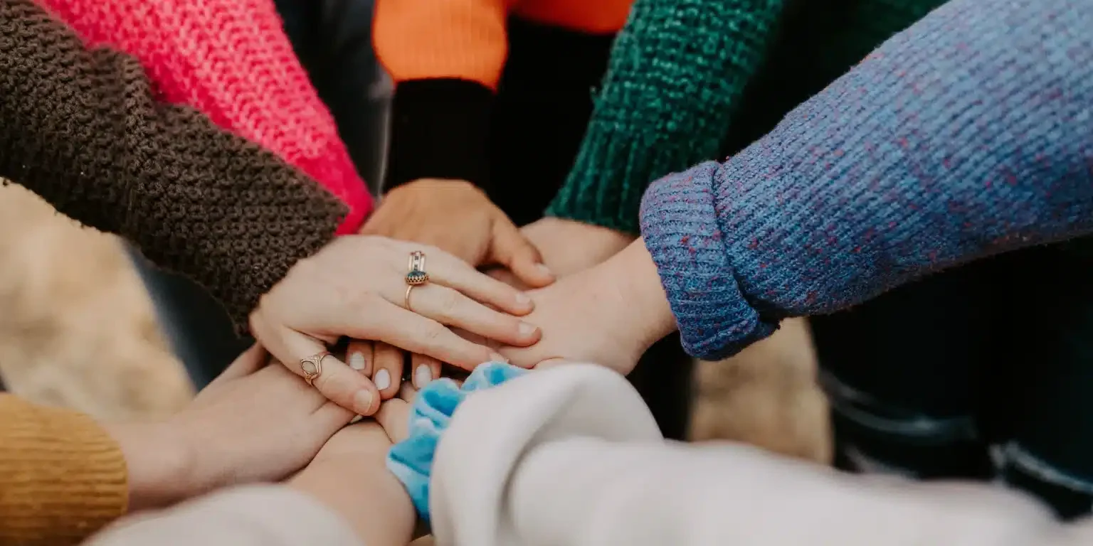 A clean group of people stands in a circle, each extending one hand into the center, layering their hands on top of one another. They are wearing colorful, knitted sweaters, suggesting unity and community teamwork.
