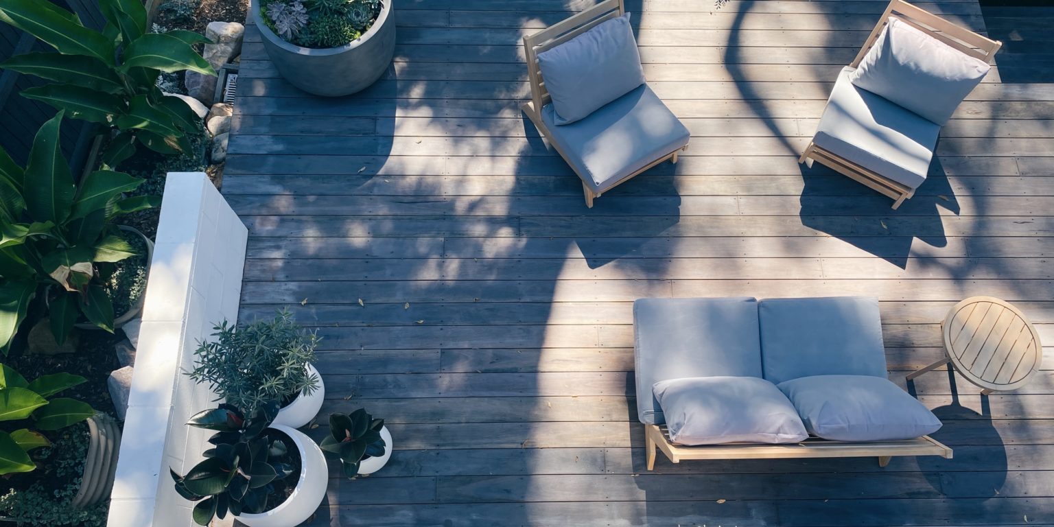 Aerial view of a clean deck featuring a wooden patio with cushioned chairs and a loveseat. The patio, perfect for the spring season, is adorned with potted plants and bathed in a mix of sunlight and shadow. Surrounding the patio is greenery, adding a natural ambiance to the cozy outdoor space.